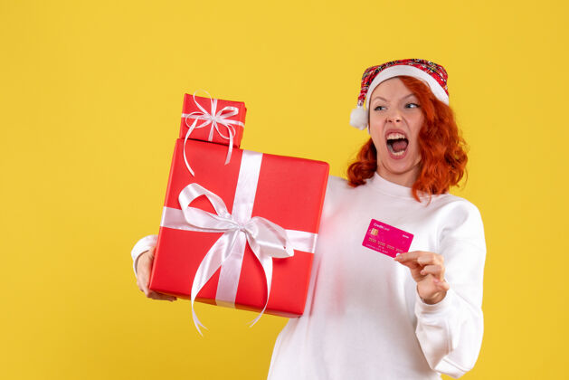 银行黄墙上年轻女子拿着圣诞礼物和银行卡的正面图圣诞节年轻的女人微笑
