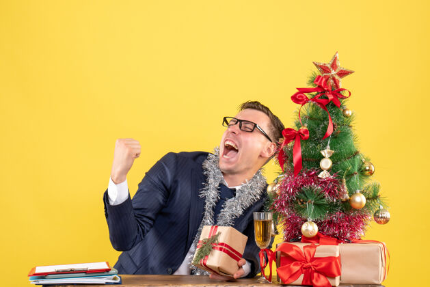 成人在圣诞树旁的桌子旁 一个欢欣鼓舞的男人展示着胜利的姿态 黄色的礼物黄色人桌子