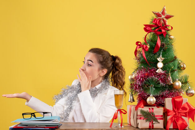 情感前视图：女医生坐在黄色背景的桌子后面 带着圣诞树和礼品盒女性圣诞节人
