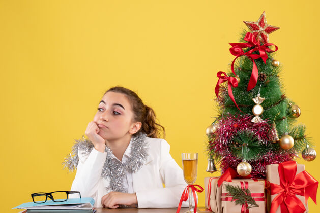 背景正面图：女医生坐在黄色背景的桌子后面 还有圣诞树和礼品盒医生前面坐着