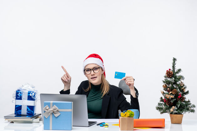 专业戴着圣诞老人帽戴着眼镜的漂亮女人坐在桌旁 拿着圣诞礼物 手里拿着银行卡 在办公室里往上看帽子圣诞老人看