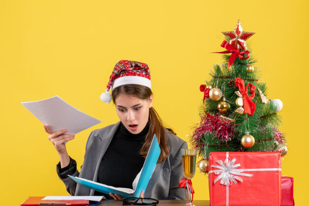 微笑前视图震惊女孩与圣诞帽坐在桌边检查文件圣诞树和礼物鸡尾酒坐着人圣诞节