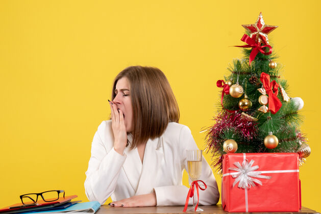 电脑正面图：女医生坐在桌子前打着哈欠 背景是黄色的圣诞树和礼盒女医生成人情绪