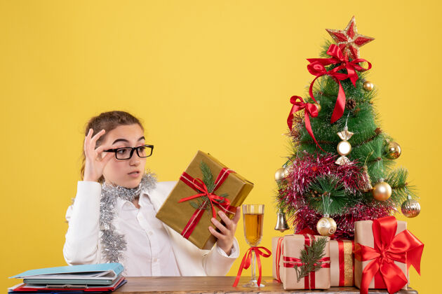 情感前视图：女医生坐在桌子后面 拿着圣诞礼物 黄色背景上的树背景新郎健康