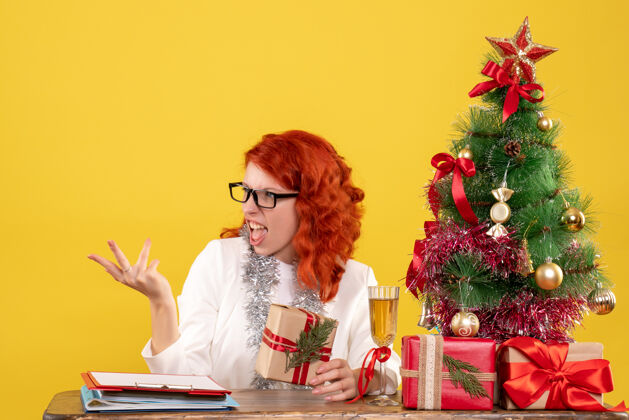背景前视图女医生坐在桌子后面 黄色背景上有圣诞礼物 还有圣诞树和礼品盒礼物女医生护士