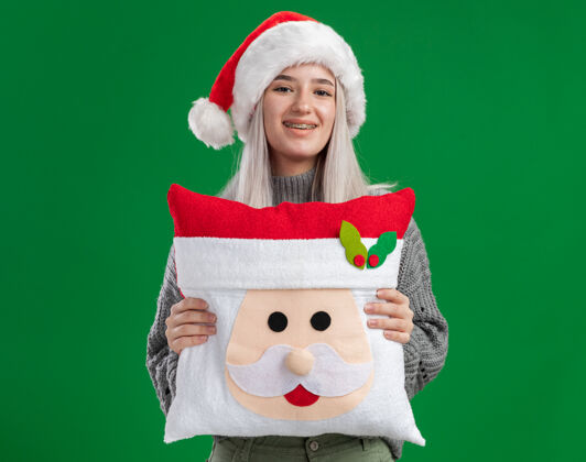 圣诞老人快乐的金发女郎 穿着冬衣 戴着圣诞帽 抱着圣诞枕头 面带微笑地看着相机 站在绿色的背景下枕头圣诞年轻