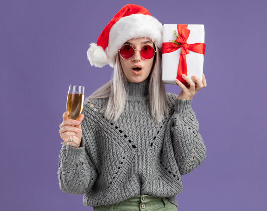 快乐年轻的金发女郎穿着冬衣 戴着圣诞帽 手里拿着礼物 手里拿着一杯香槟 站在紫色的背景下惊讶地看着镜头金发杯子礼物