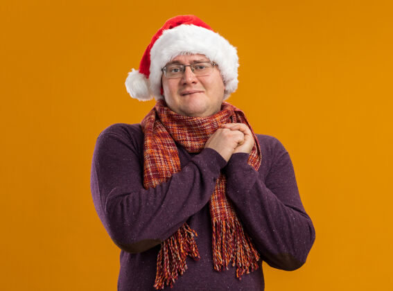 兴奋兴奋的成年男子戴着眼镜 戴着圣诞帽 脖子上围着围巾 看着镜头 手放在一起 咬着嘴唇 在橙色的背景上孤立着嘴唇一起手