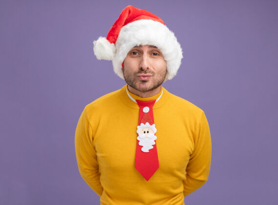 手自信的白人年轻人戴着圣诞帽 打着领带 看着摄像机 双手放在背后 做着亲吻的手势 紫色背景上 有着独立的复制空间领带亲吻看