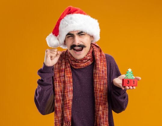 站立一个留着胡子的男人戴着圣诞老人帽 脖子上围着暖和的围巾 手里拿着玩具方块 手里攥着新年约会的拳头 站在橙色的背景上 快乐而兴奋周围新的圣诞老人