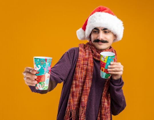 微笑快乐的胡子男人戴着圣诞老人的帽子 脖子上围着暖和的围巾 手里拿着五颜六色的纸杯 站在橙色的背景上 面带微笑地看着他们站着圣诞老人围巾