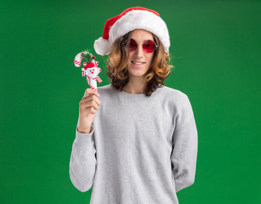 糖果快乐的年轻人戴着圣诞老人帽 戴着红眼镜 手持圣诞糖果手杖 站在绿色的背景下 面带微笑地看着相机抱着微笑帽子