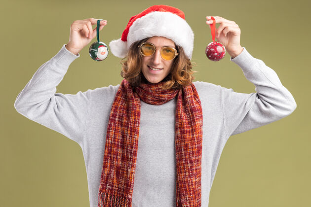 眼镜快乐的年轻人戴着圣诞老人帽 戴着黄色眼镜 脖子上围着暖和的围巾 手里拿着圣诞球 站在绿色的背景下 面带微笑地看着相机年轻帽子圣诞节