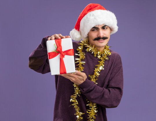 圣诞老人站在紫色的背景下 一个留着胡子的年轻人戴着圣诞老人帽子 脖子上戴着金属丝 手里拿着圣诞礼物 看上去既困惑又不高兴脖子帽子不高兴
