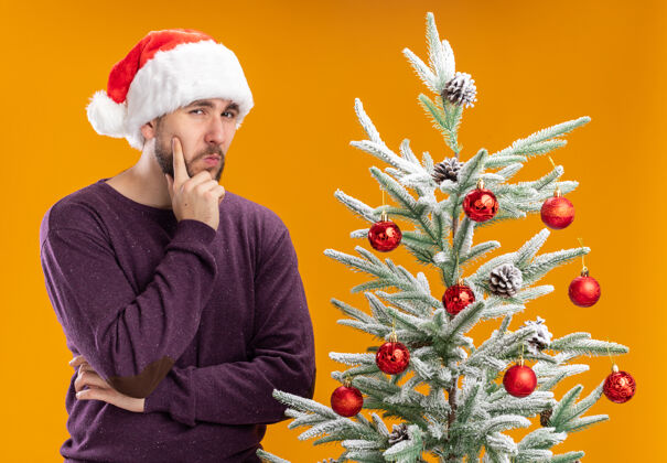 圣诞老人身穿紫色毛衣 头戴圣诞帽的年轻人 脸上带着沉思的表情 站在橙色墙上的圣诞树旁思考着表情圣诞节毛衣