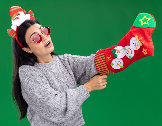 圣诞老人好奇的年轻白人女孩戴着圣诞老人的头带戴着眼镜拿着圣诞长袜看着它把手放在里面隔离在绿色的墙上里面好奇长袜