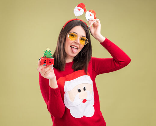 新年顽皮的年轻漂亮的白人女孩 穿着圣诞老人毛衣 头戴眼镜 手里拿着圣诞树玩具和日期圣诞日期看