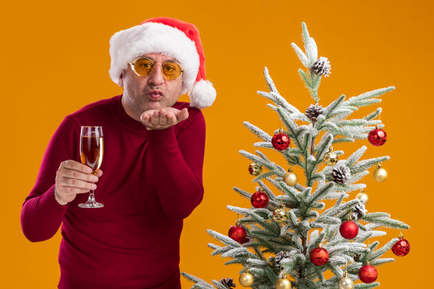 圣诞快乐的中年男人戴着圣诞老人帽 穿着深红色高领毛衣 戴着一副黄色的眼镜 手里拿着一杯香槟 站在橙色背景下的圣诞树旁亲吻中间亲吻香槟