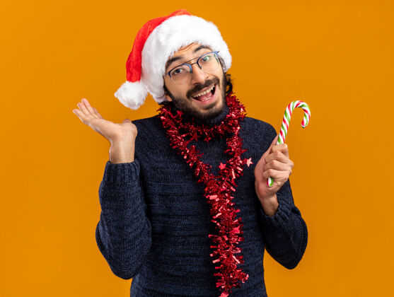 糖果微笑的年轻帅哥戴着圣诞帽 脖子上戴着花环 手里拿着圣诞糖果 手被隔离在橙色的背景上帽子帅气微笑