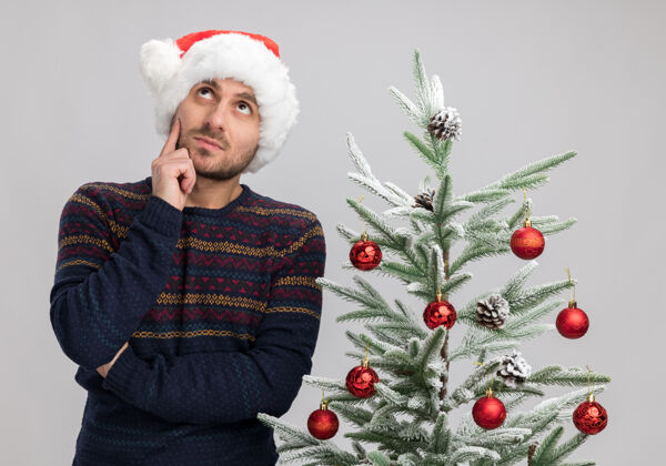 站着体贴的白人年轻人戴着圣诞帽站在圣诞树旁 抬头望着白色背景上孤立的一张动人的脸感动圣诞节帽子