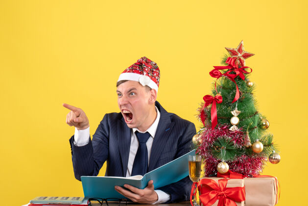 行政人员愤怒的商人对着坐在圣诞树旁桌子上的人大喊大叫 黄色墙壁上摆着礼物有人愤怒的商人树