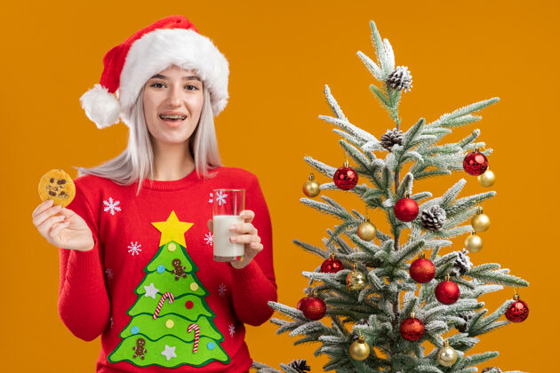 帽子穿着圣诞毛衣 戴着圣诞帽 手里拿着一杯牛奶和饼干 脸上带着微笑的金发女郎金发脸女人