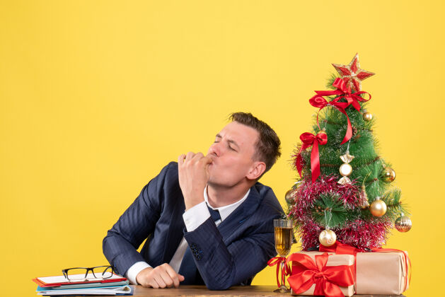 快乐极乐男子坐在圣诞树旁的桌子前做亲吻手势 黄色墙上摆着礼物男人前面人