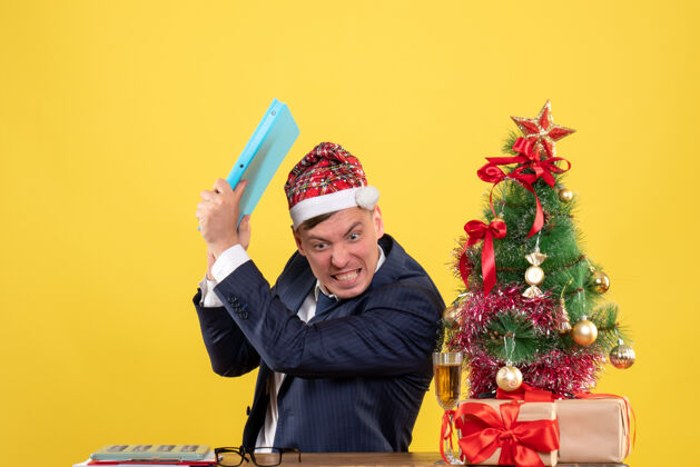 文件愤怒的商人把文件从圣诞树旁的桌子上扔掉 把礼物放在黄色的墙上工作树扔