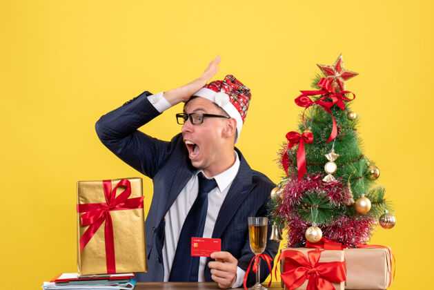 圣诞节前视图惊奇的男人把手放在额头上 坐在圣诞树旁的桌子旁 黄色的墙上摆着礼物帅哥西装男