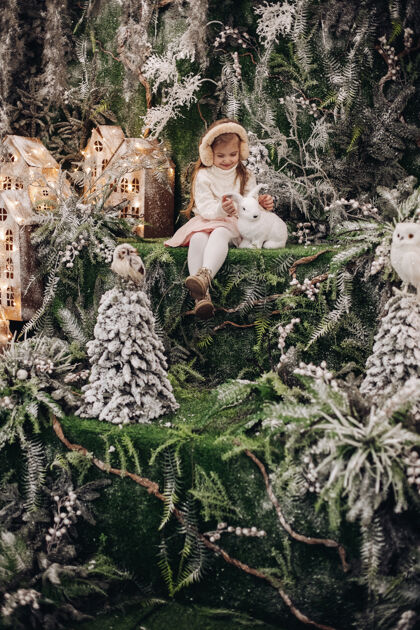 自然美丽的白种人小孩 长着金色的长发 坐在圣诞气氛中 周围有很多装饰树 还有一只小兔子快乐庆祝家