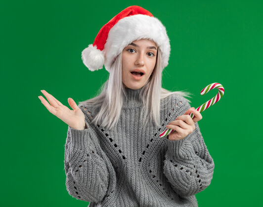 糖果年轻的金发女人穿着冬天的毛衣 戴着圣诞帽 手里拿着糖果手杖 困惑地看着摄像机 举起手臂站在绿色的背景上年轻人毛衣手杖