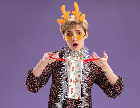 花环困惑的年轻漂亮女孩戴着驯鹿鹿角头带 脖子上戴着金箔花环 戴着眼镜 手里拿着圣诞礼包 孤零零地贴在紫色的墙上漂亮迷茫鹿角