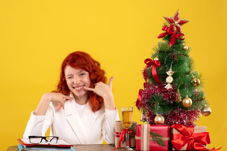 桌子女工人坐在桌子后面 手里拿着圣诞礼物 树在黄色上微笑礼物女树
