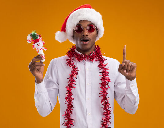 装饰品一个年轻的美国黑人男子戴着圣诞帽 戴着眼镜 脖子上戴着金箔花环 手里拿着糖果手杖 看着相机 在橙色的背景上孤立地指向上方周围帽子圣诞老人