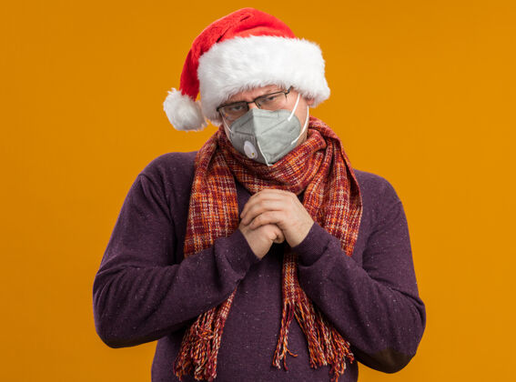 围巾狡猾的成年男子戴着防护面具眼镜 戴着圣诞老人帽 脖子上围着围巾 手放在一起看着橙色背景上孤立的摄像机脖子成人保护
