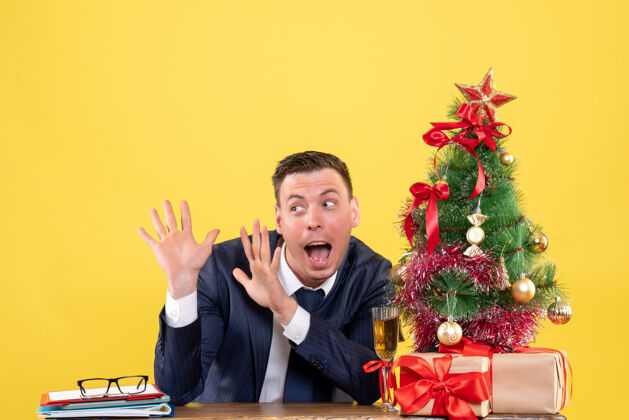 生意穿着西装的惊奇男人张开双手坐在圣诞树旁的桌子前 黄色墙上挂着礼物惊奇微笑礼物