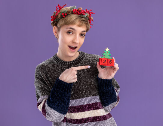 圣诞节令人印象深刻的年轻漂亮女孩戴着圣诞花环手持圣诞树玩具与日期指向它隔离在紫色墙上花环头漂亮
