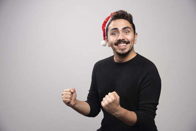 快乐戴着圣诞老人红帽子的快乐男人在灰色的墙上男孩男人成功