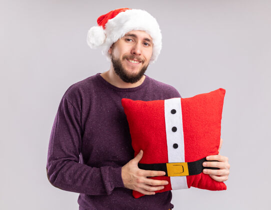 枕头快乐的年轻人穿着紫色毛衣 戴着圣诞帽 抱着滑稽的枕头 站在白色的背景下 面带微笑地看着相机年轻抱着搞笑