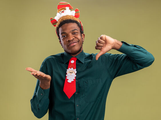 非洲一个非洲裔美国人 戴着滑稽的圣诞老人戒指 打着红色领带 看着相机 困惑地看着相机 站在绿色背景上 大拇指上下翻腾圣诞老人搞笑困惑