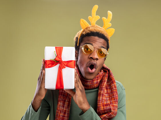 搞笑一个非洲裔美国人 戴着滑稽的边 戴着鹿角 脖子上围着围巾 手里拿着圣诞礼物 站在绿色的背景上惊讶地看着摄像机非洲角鹿
