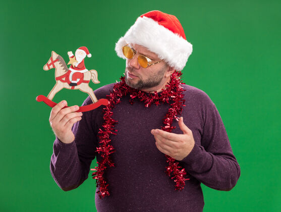 中年困惑的中年男子戴着圣诞老人帽 脖子上戴着金箔花环 戴着眼镜 看着并指着绿色背景上孤零零的摇马雕像上的圣诞老人困惑摇摆帽子