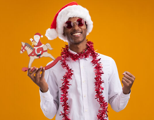 眼镜快乐的美国黑人年轻人戴着圣诞帽 戴着眼镜 脖子上戴着金箔花环 抱着摇摇木马上的圣诞老人 在橙色背景下看着相机做着“是”的手势帽子金属片马