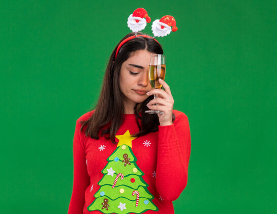 坚持失望的年轻白人女孩戴着圣诞老人的头带 拿着一杯香槟 背景是绿色的 还有复印空间失望香槟年轻