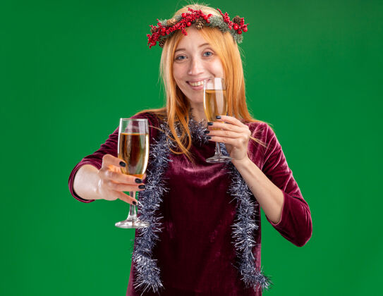 花环微笑着的年轻漂亮的女孩穿着红色的裙子 脖子上戴着花环和花环 在绿色的背景下 对着镜头举起一杯香槟抱着杯子微笑