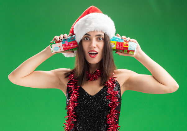 帽子惊讶的年轻漂亮女孩戴着圣诞帽 脖子上戴着花环 耳朵上戴着圣诞杯 背景是绿色的女孩圣诞节脖子