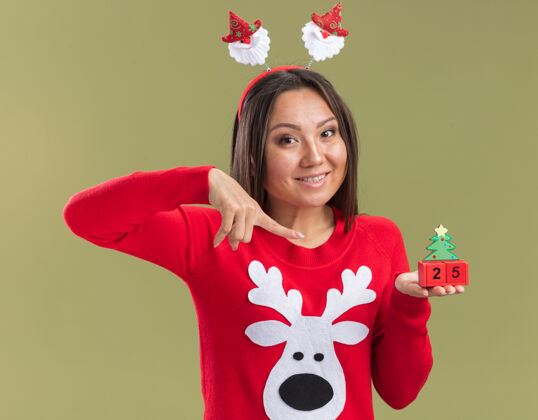 头发笑容满面的年轻亚洲女孩戴着圣诞发箍 指着橄榄绿背景上孤立的圣诞玩具圣诞节橄榄女孩