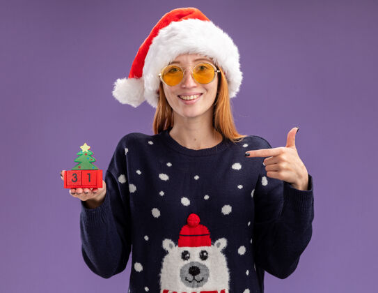 圣诞节微笑着的年轻漂亮女孩穿着圣诞毛衣 戴着圣诞帽 戴着眼镜 指着紫色背景上孤立的圣诞玩具点拿着帽子