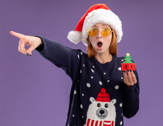 年轻惊讶的年轻漂亮女孩穿着圣诞毛衣 戴着圣诞帽 戴着眼镜 拿着圣诞玩具点在一旁 紫色背景上孤立着点圣诞节毛衣