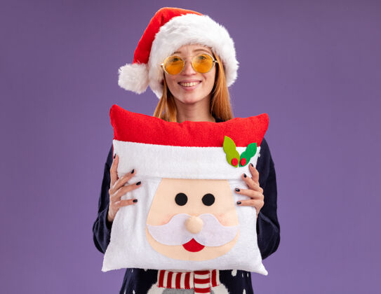 女孩微笑着的年轻漂亮女孩穿着圣诞毛衣 戴着圣诞帽 戴着眼镜 抱着一个紫色背景的圣诞枕头眼镜年轻人圣诞节